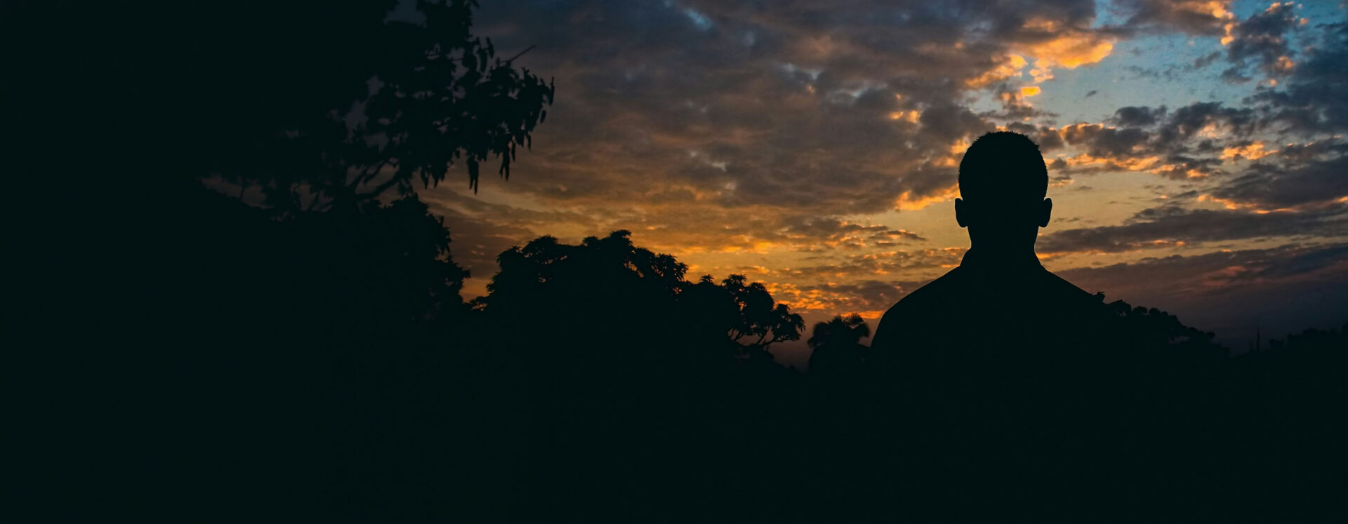 person standing alone beneath a dramatic sunset sky, observing the wider landscape from a position slightly apart, symbolizing independent perspective and insight