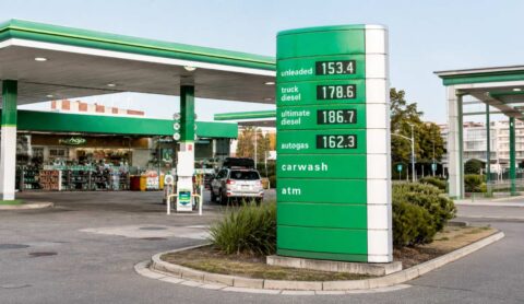 Gas station with a green-and-white canopy and a tall green price sign showing fuel types and prices, plus a shop in the background.