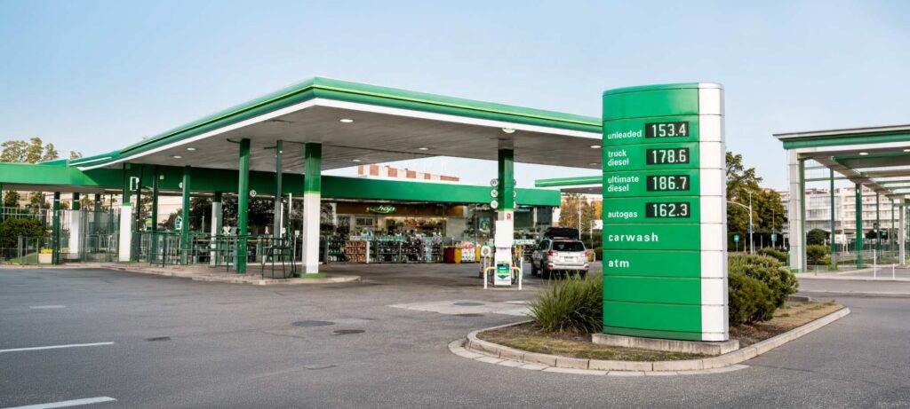 Gas station with a green-and-white canopy and a tall green price sign showing fuel types and prices, plus a shop in the background. - IMD Business School - IMD Business School