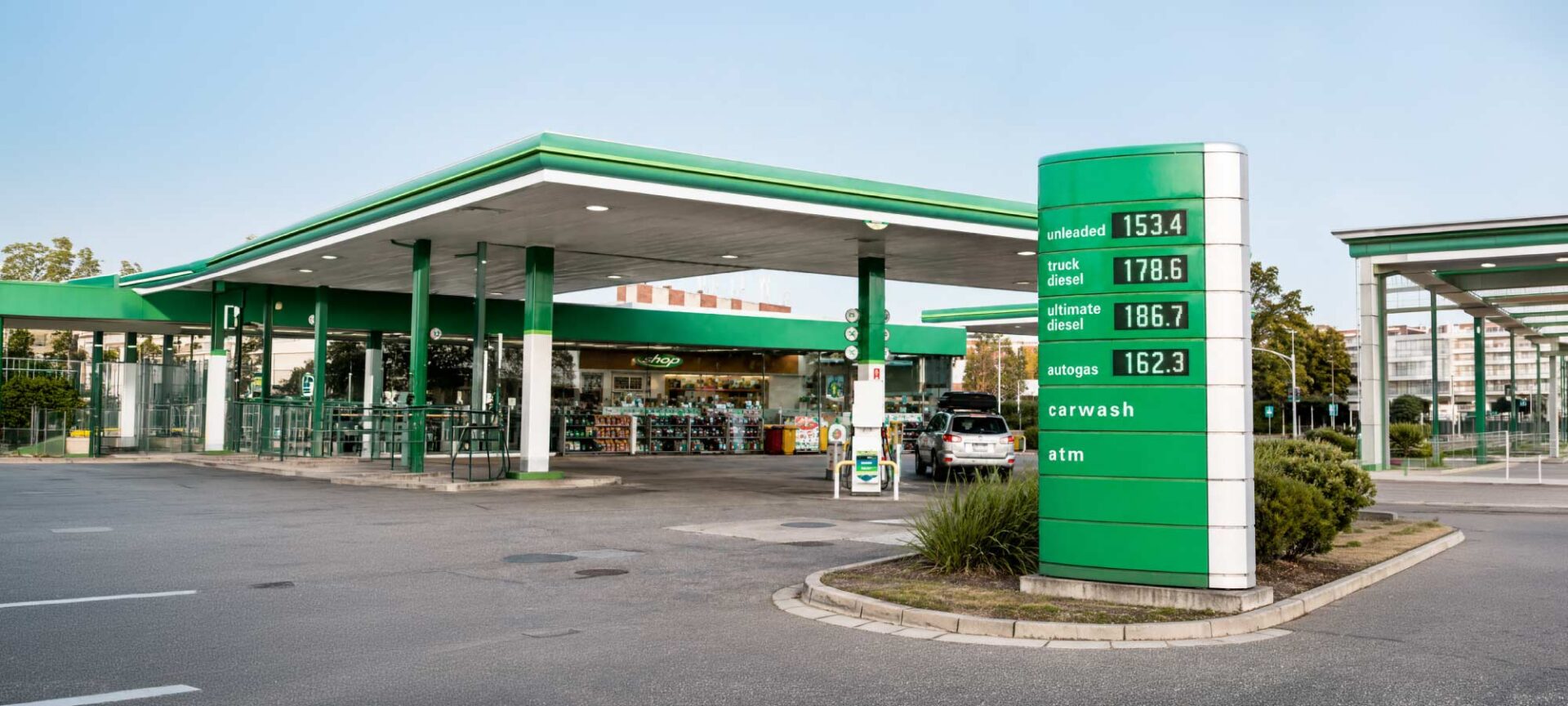 Gas station with a green-and-white canopy and a tall green price sign showing fuel types and prices, plus a shop in the background.