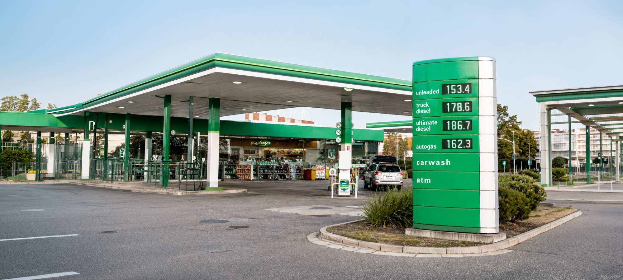 Gas station with a green-and-white canopy and a tall green price sign showing fuel types and prices, plus a shop in the background.