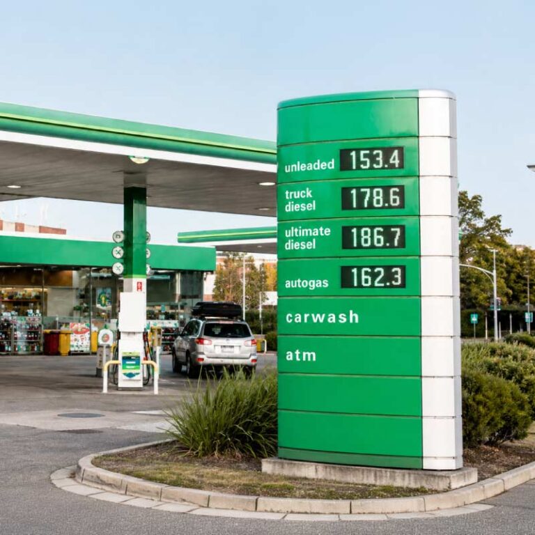 Gas station with a tall green price board listing fuel types and prices (unleaded 153.4, truck diesel 178.6, ultimate diesel 186.7, autogas 162.3) plus carwash and ATM; a car is parked near pumps under a green canopy.