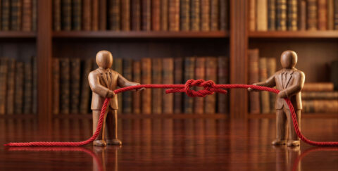 mannequin figures standing on a dark wooden table in front of bookshelves, engaged in a tug-of-war with a red rope tied in a knot, symbolizing tension and the rift between leadership and employees in a toxic workplace