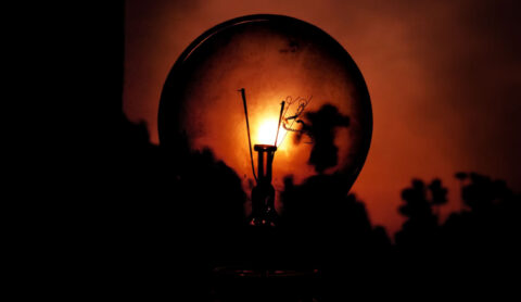 Close-up of a glowing incandescent light bulb casting a warm orange glow against a dark background, filament visible inside the bulb.