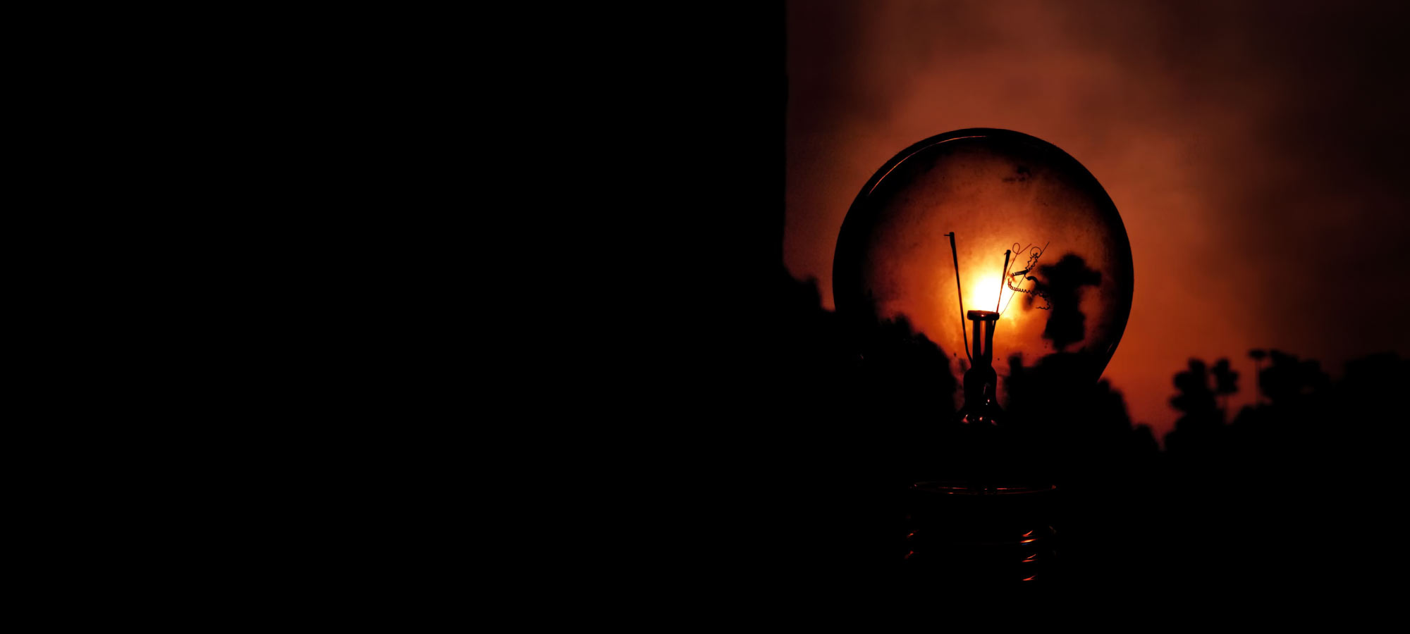 Close-up of a glowing incandescent light bulb casting a warm orange glow against a dark background, filament visible inside the bulb.