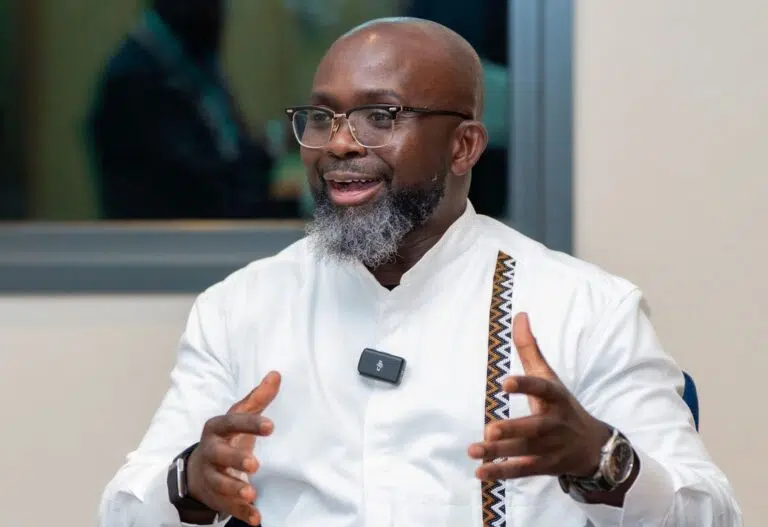 Isaac Fokuo speaking animatedly during an interview, gesturing with his hands while wearing a white shirt and glasses. - IMD Business School